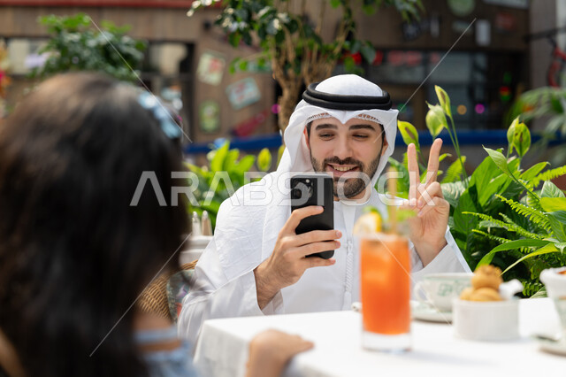 مفهوم الألفة والمحبة بين أفراد الأسرة، الاحتفاظ بالذكريات السعيدة، قضاء وقت ممتع في الخارج، صورة مقربة لرجل عربي خليجي اماراتي يرتدي الكندورة والغترة يجلس باحدى الكافيهات يرفع يده بايماءات النصر يقوم بالتقاط صورة تذكارية لابنته باستخدام الهاتف المحمول