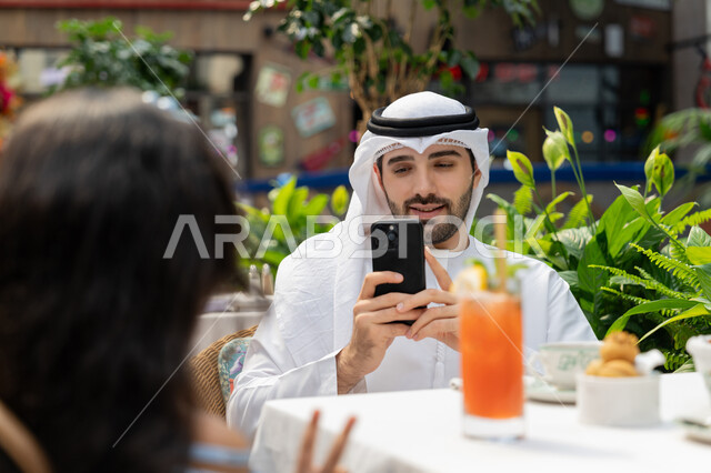 قضاء وقت ممتع في الخارج، الاحتفاظ بالذكريات السعيدة، مفهوم الألفة والمحبة بين أفراد الأسرة, أجواء اسرية مليئة بالحب، صورة مقربة لرجل عربي خليجي اماراتي يرتدي الكندورة والغترة يجلس باحدى الكافيهات يقوم بالتقاط صورة تذكارية لابنته باستخدام الهاتف المحمول