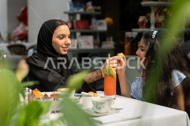 Gestures of joy and happiness, a happy family atmosphere full of love, entertainment and fun during holidays, spending time outdoors, family entertainment activities and events, the concept of intimacy and love between family members, a close-up photo of an Arab Gulf Emirati father sitting in a cafe offering a cup of juice to his daughter