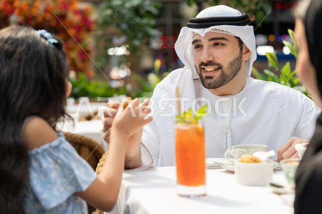 قضاء الوقت في الخارج، التسلية والترفيه بالعطلة، أجواء اسرية سعيدة مليئة بالحب، انشطة وفعاليات ترفيهية عائلية، مفهوم الألفة والمحبة بين أفراد الأسرة، صورة مقربة لاب عربي خليجي اماراتي يجلس في المقهى يقوم بتقديم قطعة من الحلوى لابنته، تناول العصير والمشروبات الباردة