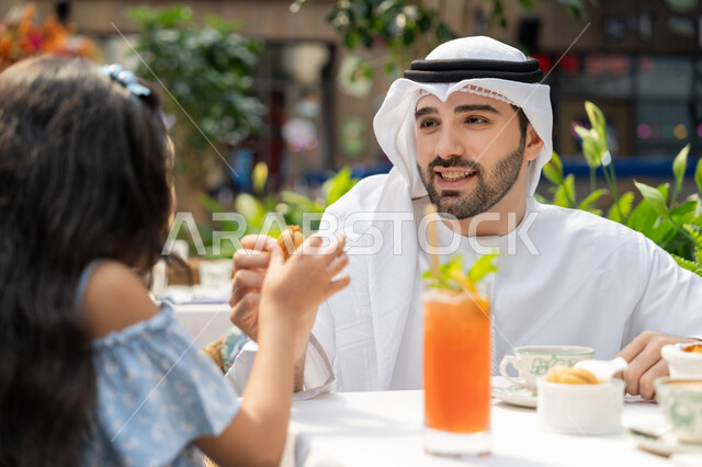 Holiday entertainment and fun, happy family atmosphere full of love, spending time outdoors, family entertainment activities and events, the concept of intimacy and love between family members, a close-up photo of an Arab Gulf Emirati father sitting in a cafe offering a piece of candy to his daughter, drinking juice and cold drinks