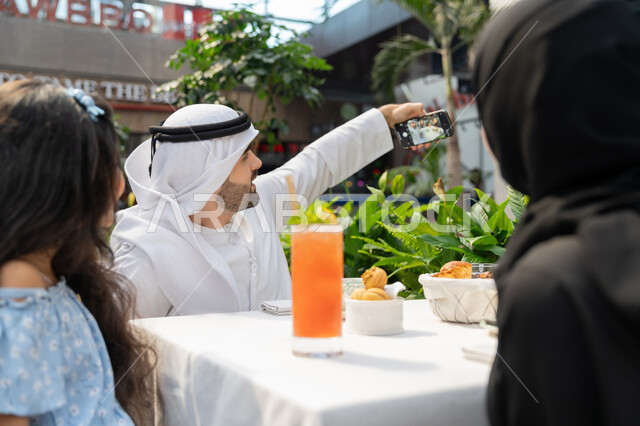 الاحتفاظ بالذكريات السعيدة، مفهوم الألفة والمحبة بين أفراد الأسرة, قضاء وقت ممتع في الخارج، صورة مقربة لرجل عربي خليجي اماراتي يرتدي الكندورة والغترة يجلس باحدى الكافيهات يقوم بالتقاط صورة تذكارية برفقة عائلته باسخدام الهاتف المحمول