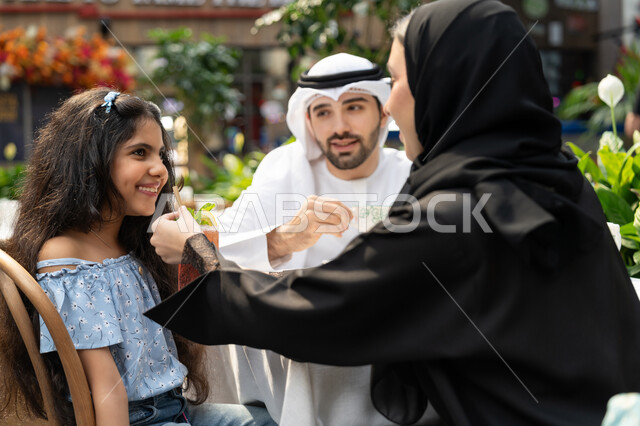 The concept of intimacy and love between family members, spending time outdoors, gestures of pleasure and happiness, entertainment and recreation during holidays, a happy family atmosphere full of love, family entertainment activities and events, a close-up photo from the back of an Arab Gulf Emirati mother sitting in a cafe offering sweets to her daughter