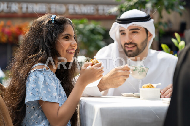 استغلال وقت الفراغ في التسلية مع الاسرة، قضاء أوقات ممتعة في الخارج، أجواء عائلية سعيدة مليئة بالحب والحنان، صورة مقربة من الجانب لفتاة عربية خليجية اماراتية تجلس في احدى الكافيهات برفقة والدها تنظر لمكان ما بايماءات السرور والسعادة