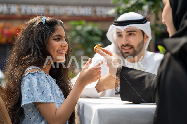 Spending time outdoors, gestures of pleasure and happiness, entertainment and leisure during the holidays, a happy family atmosphere full of love, family entertainment activities and events, the concept of intimacy and love between family members, a close-up photo from the back of an Arab Gulf Emirati mother sitting in a cafe offering sweets to her daughter