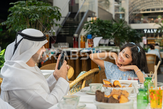 أجواء اسرية سعيدة مليئة بالحب والحنان، مفهوم الألفة والمحبة بين أفراد الأسرة, قضاء وقت ممتع في الخارج، صورة مقربة من الجانب لرجل عربي خليجي اماراتي يرتدي الكندورة والغترة يجلس باحدى الكافيهات يقوم بالتقاط صورة تذكارية لابنته باستخدام الهاتف المحمول