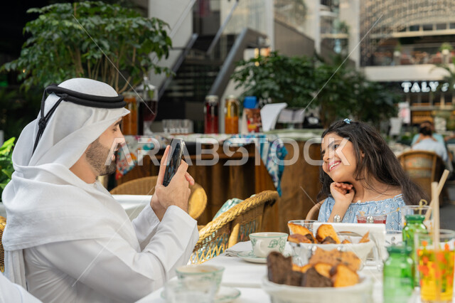 The concept of intimacy and love between family members, a happy family atmosphere full of love and tenderness, spending a pleasant time outside, a close-up photo from the side of an Emirati Gulf Arab man wearing a kandura and a ghutra sitting in a café taking a souvenir photo of his daughter using a mobile phone