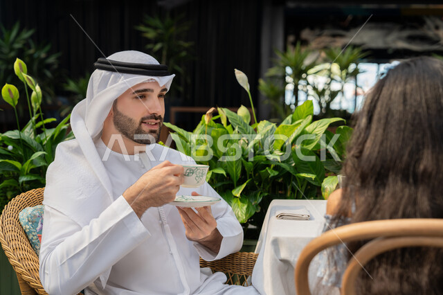 حوار ونقاش عائلي، الاستمتاع بتناول المشروبات الساخنة، قضاء وقت ممتع في الخارج، مفهوم الألفة والمحبة بين أفراد الأسرة, صورة مقربة لرجل عربي خليجي اماراتي يرتدي الكندورة والغترة يجلس برفقة ابنته في احدى الكافيهات يقوم بشرب القهوة