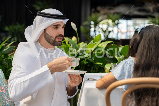 الاستمتاع بتناول المشروبات الساخنة، حوار ونقاش عائلي، قضاء وقت ممتع في الخارج، مفهوم الألفة والمحبة بين أفراد الأسرة, صورة مقربة لرجل عربي خليجي اماراتي يرتدي الكندورة والغترة يجلس برفقة ابنته في احدى الكافيهات يقوم بشرب القهوة