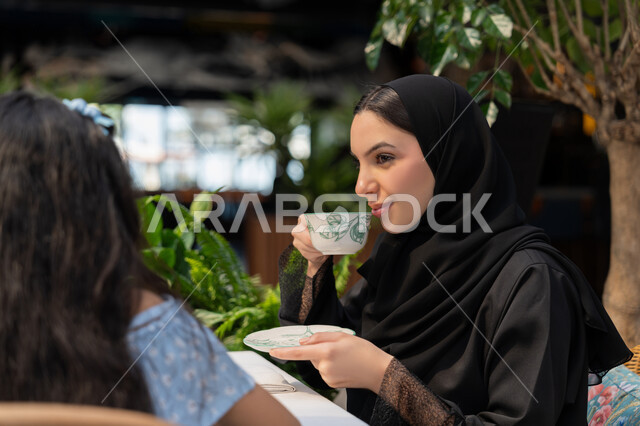 الاستمتاع بتناول المشروبات الساخنة، أجواء عائلية سعيدة مليئة بالحب والحنان، مفهوم الألفة والمحبة بين أفراد الأسرة, صورة مقربة لام عربية خليجية اماراتية محجبة ترتدي العباءة تقوم بقضاء وقت ممتع في الخارج برفقة ابنتها، امرأة سعودية تقوم بشرب القهوة في احدى الكافيهات