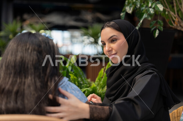 استغلال وقت الفراغ في التسلية، أجواء عائلية سعيدة مليئة بالحب، قضاء أوقات ممتعة في الخارج، مفهوم الألفة والمحبة بين أفراد الأسرة, صورة مقربة لام عربية خليجية اماراتية محجبة ترتدي العباءة تقوم بمداعمة ابنتها، صورة من الخلف لفتاة تجلس مع امها في احدى الكافيهات