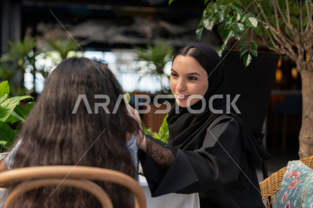أجواء عائلية سعيدة مليئة بالحب، قضاء أوقات ممتعة في الخارج، مفهوم الألفة والمحبة بين أفراد الأسرة, صورة مقربة لام عربية خليجية اماراتية محجبة ترتدي العباءة تقوم بمداعمة ابنتها، صورة من الخلف لفتاة تجلس مع امها في احدى الكافيهات، استغلال وقت الفراغ في التسلية