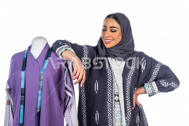 بورتريه لمصممة أزياء خليجية سعودية مبتسمة وفخورة بعد الإنتهاء من تصميمها المعلق على المانيكان، مصممة أزياء مبتكرة، ورشة عمل تصميم ازياء، أدوات الخياطة، مهن وأعمال فن الخياطة، فنون ومهارات تصميم وتفصيل الملابس النسائية، مفهوم عالم الأزياء والموضة