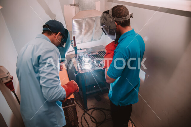 تجهيز وتوصيل الحديد بمعدات وأدوات لحامية, مهن وأعمال سعوديه، المهارة في اتقان العمل في مصنع الحداده, صورة من الخلف لمهندسين صناعيين عربيين خليجيين سعوديين يرتدون قناع الحماية والقفازات يقومون بلحام المعدن