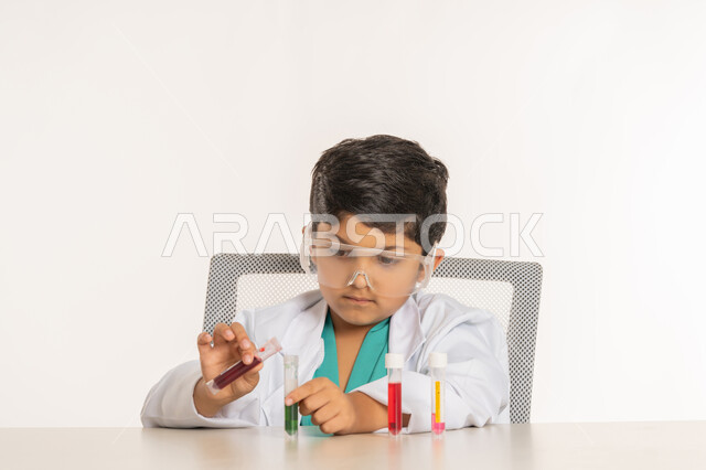 Scientific research and experiments, choosing and determining the future from a young age, the profession of a bio-laboratory specialist, a close-up portrait of an Arab Gulf Emirati boy wearing a white coat sitting in front of a table wearing protective glasses performing chemical reactions, a Saudi boy using test tubes, white background