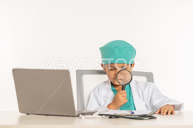 توضيح وتحسين رؤية، أدوات بصرية للتدقيق والتركيز، مفهوم الطب والرعاية الصحية، استخدام الاجهزة التقنية، بورتريه مقرب لولد عربي خليجي اماراتي يرتدي المعطف الأبيض يضع السماعة الطبية يجلس خلف المكتب يقوم بمراجعة سجل المرضى، صبي سعودي يحمل بيده عدسة مكبرة، خلفية بيضاء