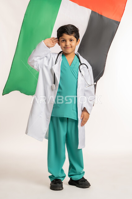 الاحتفال بالأعياد والمناسبات الوطنية, العمل في القطاع الطبي، رفع راية الامارات العربية المتحدة عاليا، ذكرى اليوم الوطني الإماراتي 2 ديسمبر, بورتريه لولد عربي خليجي إماراتي يرتدي المعطف يضع السماعات الطبية يقوم باداء التحية العلم, صورة لكامل الجسم، خلفية بيضاء