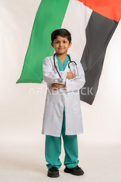 رفع علم الامارات عاليا، العمل في القطاع الطبي، ذكرى اليوم الوطني الإماراتي 2 ديسمبر, بورتريه لولد عربي خليجي إماراتي يرتدي المعطف الابيض يضع السماعات الطبية يقف بايدي متقاطعة ينظرالى الكاميرا بايماءات الفخر والاعتزاز, صورة لكامل الجسم، خلفية بيضاء