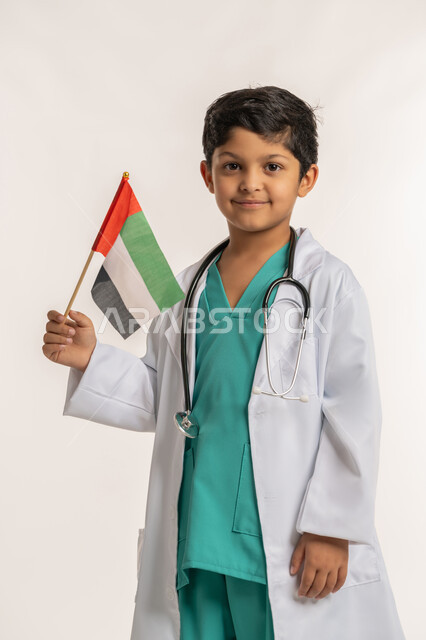 العمل في القطاع الطبي، النظر إلى الكاميرا بتعابير الفخر بالانتماء للبلاد, الاحتفال بالأعياد والمناسبات الوطنية, ذكرى اليوم الوطني الإماراتي 2 ديسمبر, بورتريه لولد عربي خليجي إماراتي يرتدي المعطف يضع السماعات الطبية يحمل علم الإمارات, خلفية بيضاء