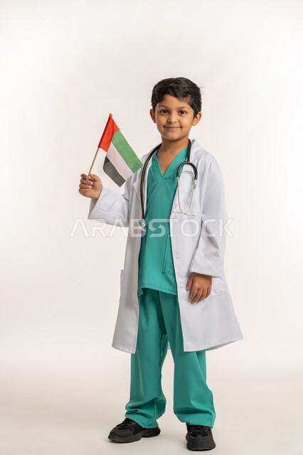 النظر إلى الكاميرا بتعابير الفخر بالانتماء للبلاد, العمل في القطاع الطبي، الاحتفال بالأعياد والمناسبات الوطنية, ذكرى اليوم الوطني الإماراتي 2 ديسمبر, بورتريه لولد عربي خليجي إماراتي يرتدي المعطف يضع السماعات الطبية يحمل علم الإمارات, الجسم بالطول الكامل، خلفية بيضاء