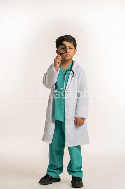 توضيح وتحسين رؤية، أدوات بصرية للتدقيق والتركيز، مفهوم الطب والرعاية الصحية، تحديد مهنة المستقبل، بورتريه لولد عربي خليجي اماراتي يرتدي المعطف الأبيض يضع السماعة الطبية يحلم بالعمل كطبيب، صبي سعودي يحمل بيده عدسة مكبرة، صورة لكامل الجسم، خلفية بيضاء
