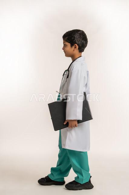 مفهوم الطب والرعاية الصحية، تحديد مهنة المستقبل، الهدف والطموح المستقبلي بالعمل في العيادات والمشافي، بورتريه من الجانب لولد عربي خليجي اماراتي يرتدي المعطف الأبيض يضع السماعة الطبية يحلم بالعمل كطبيب، صبي سعودي يحمل بيده سجل المرضى، صورة لكامل الجسم، خلفية بيضاء