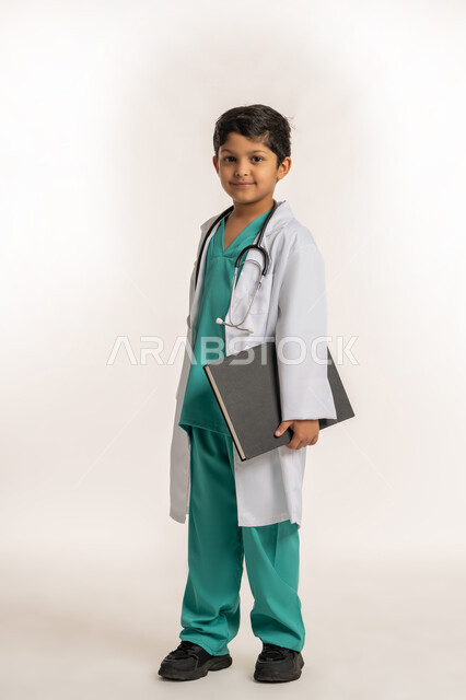 الهدف والطموح المستقبلي بالعمل في العيادات والمشافي، مفهوم الطب والرعاية الصحية، تحديد مهنة المستقبل، بورتريه لولد عربي خليجي اماراتي يرتدي المعطف الأبيض يضع السماعة الطبية يحلم بالعمل كطبيب، صبي سعودي يحمل بيده سجل المرضى، صورة لكامل الجسم، خلفية بيضاء