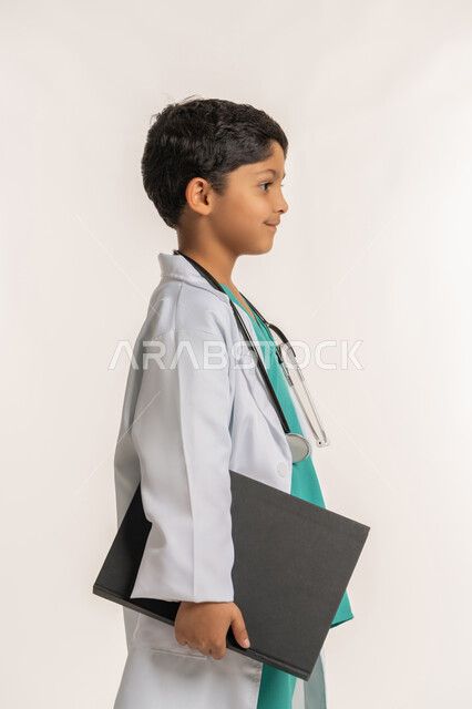 الهدف والطموح المستقبلي بالعمل في العيادات والمشافي، مفهوم الطب والرعاية الصحية، تحديد مهنة المستقبل، بورتريه من الجانب لولد عربي خليجي اماراتي يرتدي المعطف الأبيض يضع السماعة الطبية يحلم بالعمل كطبيب، صبي سعودي يحمل بيده سجل المرضى، خلفية بيضاء