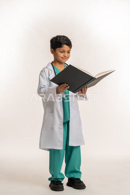 الهدف والطموح المستقبلي بالعمل في العيادات والمشافي، مفهوم الطب والرعاية الصحية، بورتريه لولد عربي خليجي اماراتي يرتدي المعطف الأبيض يضع السماعة الطبية يحمل بيده سجل المرضى، فتى سعودي يقوم بالقراءة بايماءات التركيز، صورة لكامل الجسم، خلفية بيضاء