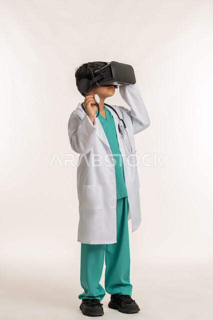 تحديد المهنة المستقبلية منذ الصغر، مفهوم الطب والرعاية الصحية، الاندماج مع العالم الخيالي، بورتريه لولد عربي خليجي اماراتي يرتدي المعطف الأبيض يضع نظارة الفي ار يقوم بالتفاعل مع الواقع الافتراضي، صبي سعودي يمسك بيده السماعة الطبية، صورة لكامل الجسم، خلفية بيضاء