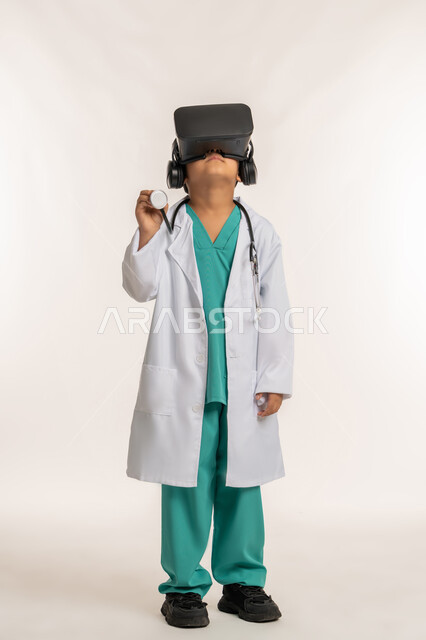 تحديد المهنة المستقبلية منذ الصغر، مفهوم الطب والرعاية الصحية، الاندماج مع العالم الخيالي، بورتريه لولد عربي خليجي اماراتي يرتدي المعطف الأبيض يضع نظارة الفي ار يقوم بالتفاعل مع الواقع الافتراضي، صبي سعودي يمسك بيده السماعة الطبية، صورة لكامل الجسم، خلفية بيضاء