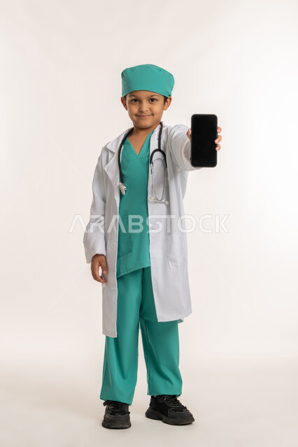 تحديد المهنة المستقبلية منذ الصغر، تعابير الشغف لتحقيق حلم الطفوله، تقديم الرعاية والخدمات الصحية، بورتريه لولد عربي خليجي اماراتي يرتدي المعطف الأبيض يضع السماعات الطبية يحمل بيده الهاتف المحمول، فتى سعودي يقوم بعرض شاشة سوداء فارغة، صورة لكامل الجسم، خلفية بيضاء