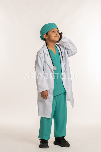 اختيار وتحديد المهنة المستقبلية منذ الصغر، تقديم الرعاية والخدمات الصحية، بورتريه لولد عربي خليجي اماراتي يرتدي المعطف الأبيض يضع السماعات الطبية يحمل بيده الهاتف المحمول، فتى سعودي يقوم باجراء مكالمة هاتفية، صورة لكامل الجسم، خلفية بيضاء