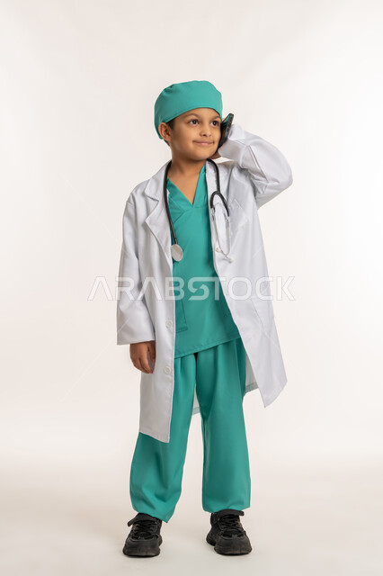 اختيار وتحديد المهنة المستقبلية منذ الصغر، تقديم الرعاية والخدمات الصحية، بورتريه لولد عربي خليجي اماراتي يرتدي المعطف الأبيض يضع السماعات الطبية يحمل بيده الهاتف المحمول، فتى سعودي يقوم باجراء مكالمة هاتفية، صورة لكامل الجسم، خلفية بيضاء