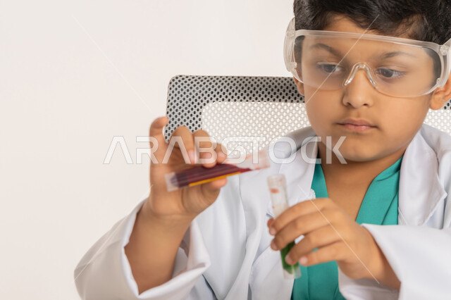 التجارب والأبحاث العلمية، مهنة اخصائي مختبرات حيوية، اختيار وتحديد المستقبل منذ الصغر، بورتريه مقرب لولد عربي خليجي اماراتي يرتدي المعطف الأبيض يجلس امام الطاولة يضع نظارات واقية يقوم باجراء التفاعلات الكيميائية، فتى سعودي يستخدام أنابيب الأختبار، خلفية بيضاء