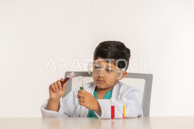 اختيار وتحديد المستقبل منذ الصغر، الأبحاث والتجارب العلمية، مهنة اخصائي مختبرات حيوية، بورتريه مقرب لولد عربي خليجي اماراتي يرتدي المعطف الأبيض يجلس امام الطاولة يضع نظارات واقية يقوم باجراء التفاعلات الكيميائية، فتى سعودي يستخدام أنابيب الأختبار، خلفية بيضاء