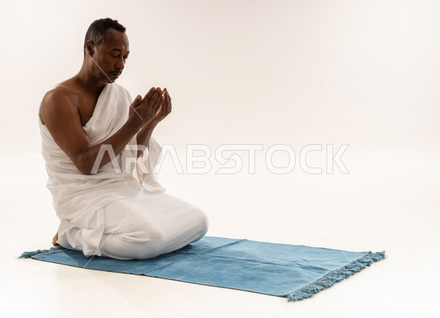 الركن الثاني من أركان الإسلام، سيد عماني يقوم بأداء مناسك الحج والعمرة ، مفهوم العبادة و التقرب الى الله، أهمية الالتزام بالصلاة على وقتها, بورتريه لرجل عربي سعودي خليجي يرتدي ملابس الاحرام يرفع يديه للأعلى بايماءات الدعاء ويجلس على سجادة الصلاة، خلفية بيضاء