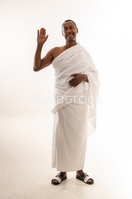 النظر إلى الكاميرا بايماءات السعادة والسرور، حاج عماني يقوم بأداء مناسك الحج والعمرة، بورتريه لرجل عربي سعودي خليجي مبتسم يرتدي ملابس الاحرام ويرفع يده للأعلى بتعابير إلقاء التحية والسلام، الجسم بالطول الكامل، خلفية بيضاء