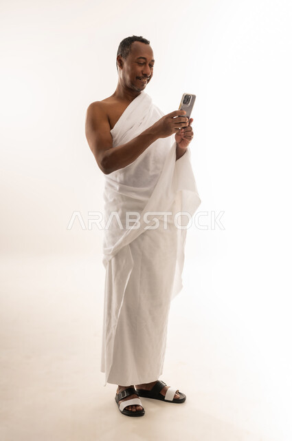 استخدام تطبيقات الارشادات لأداء مناسك الحج والعمرة، التواصل مع العائلة والأصدقاء، تطبيق الادعية والاذكار، بورتريه لحاج مسلم خليجي عربي سعودي مبتسم يرتدي ملابس الإحرام يستخدم الهاتف المحمول، رجل عماني يستخدم جهاز تقني حديث ومتطور، الجسم بالطول الكامل، خلفية بيضاء
