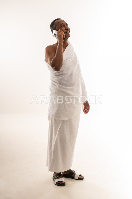 استخدام جهاز تقني حديث ومتطور، التواصل مع العائلة والأصدقاء، رجل عماني يقوم بزيارة بيت الله الحرام في مكة المكرمة، بورتريه لحاج عربي خليجي سعودي مبتسم يرتدي ملابس الاحرام يقوم بإجراء مكالمة صوتية عبر الهاتف المحمول , الجسم بالطول الكامل، خلفية بيضاء