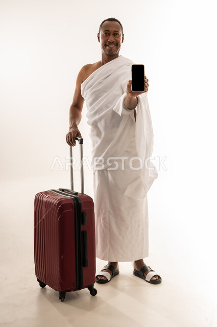 الذهاب لأداء فريضة الحج والعمرة، الاستعداد للتوجه و الانطلاق لزيارة بيت الله الحرام،  بورتريه لسيد خليجي عربي سعودي مبتسم يرتدي ملابس الاحرام ويمسك بيده حقيبة السفر، رجل عماني يقوم بعرض شاشة سوداء فارغة عبر الهاتف المحمول، الجسم بالطول الكامل، خلفية بيضاء