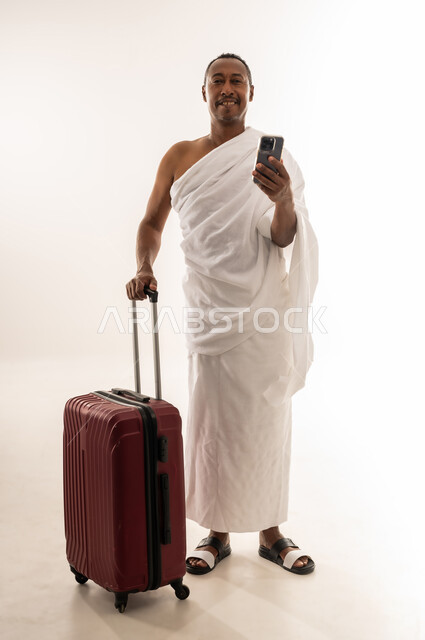 الاستعداد للتوجه والانطلاق لزيارة بيت الله الحرام، أداء فريضة الحج والعمرة، بورتريه لسيد خليجي عربي سعودي مبتسم يرتدي ملابس الاحرام يحمل بيده الهاتف المحمول وبجانبه حقيبة السفر، رجل عماني يقوم بالتواصل مع الاهل والاصدقاء، الجسم بالطول الكامل، خلفية بيضاء