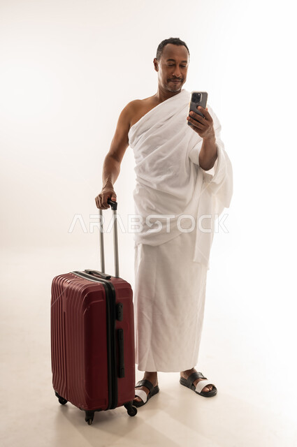 أداء فريضة الحج والعمرة، الاستعداد للتوجه والانطلاق لزيارة بيت الله الحرام، بورتريه لسيد خليجي عربي سعودي مبتسم يرتدي ملابس الاحرام يحمل بيده الهاتف المحمول وبجانبه حقيبة السفر، رجل عماني يقوم بالتواصل مع الاهل والاصدقاء، الجسم بالطول الكامل، خلفية بيضاء