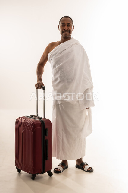 النظر الى الكاميرا بايماءات الحماس للوصول لمكة المكرمة، الاستعداد للتوجه و الانطلاق لزيارة بيت الله الحرام، بورتريه لسيد خليجي عربي سعودي مبتسم يرتدي ملابس الاحرام ويمسك بيده حقيبة السفر، رجل عماني يقوم بإداء فريضة الحج والعمرة، الجسم بالطول الكامل، خلفية بيضاء