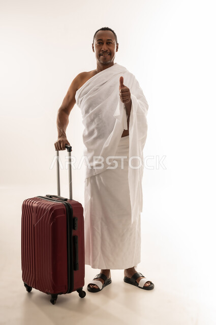 رفع اصبع الابهام بايماءات الجودة والامتياز، الاستعداد للتوجه و الانطلاق لزيارة بيت الله الحرام، بورتريه لسيد خليجي عربي سعودي مبتسم يرتدي ملابس الاحرام ويمسك بيده حقيبة السفر، رجل عماني يقوم بإداء فريضة الحج والعمرة، الجسم بالطول الكامل، خلفية بيضاء