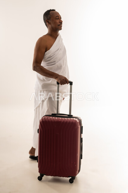 المشي و الانطلاق لزيارة بيت الله الحرام، الذهاب لأداء فريضة الحج والعمرة، بورتريه لسيد خليجي عربي سعودي مبتسم يرتدي ملابس الاحرام ويمسك بيده حقيبة السفر، رجل عماني يقف بايماءات السعادة والحماس للوصول لمكة المكرمة،  الجسم بالطول الكامل، خلفية بيضاء