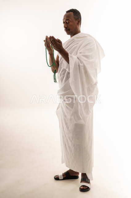 اداء مناسك الحج والعمرة، قراءة الأذكار والأدعية، التسبيح والتهليل وذكر الله تعالى، بورتريه لرجل عربي سعودي خليجي يرتدي لباس الإحرام يمسك بيده السبحة، مفهوم العبادة والتقرب إلى الله، سيد عماني يرفع يديه للأعلى بإيماءات الدعاء، الجسم بالطول الكامل، خلفية بيضاء