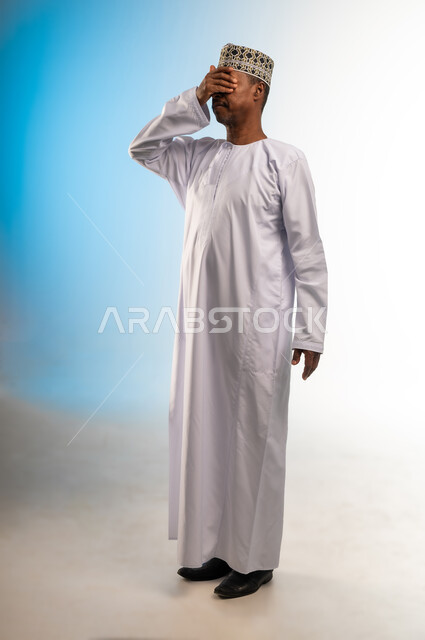 ضعف النظر وتشوش الرؤية، التعرض لأذية في الشبكية، وضع اليد على العين وتغطيها، التهاب واحمرار العيون والحاجة لزيارة الطبيب، بورتريه لرجل عربي خليجي عماني يرتدي الدشداشة والكمة يعاني من مشاكل صحية في عينه، الجسم بالطول الكامل، خلفية ملونة