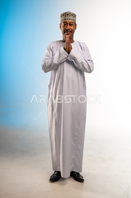 الطلب بترجي وتذلل وخضوع، مفهوم الشكر والامتنان، الوقوف بإيماءات الدعاء والتوسل والاسترحام، بورتريه لرجل عربي خليجي عماني مبتسم يرتدي الدشداشة و الكمه ينظر الى الكاميرا يرفع يديه بخشوع واستعطاف، طلب العون والرغبة في تحقيق الأمنيات، الجسم بالطول الكامل، خلفية ملونة
