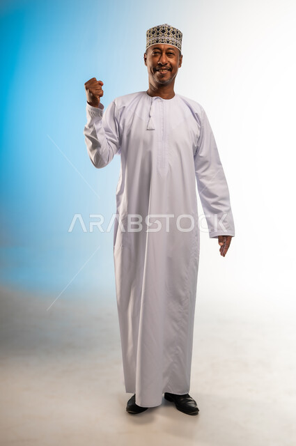 الشعور بالسعادة والقوة، مفهوم الانجاز وتحقيق الاهداف، بورتريه لرجل عربي خليجي عماني مبتسم يرتدي الدشداشة و الكمه ينظر الى الكاميرا يرفع قبضة يده للاعلى بإيماءات الفرح والسرور, الانتصار والفوز والنجاح، الجسم بالطول الكامل، خلفية ملونة